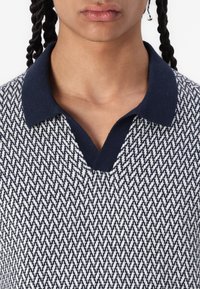 Sweter polo z dzianiny, wyróżniający się wzorem w jodełkę w granatowo-białych odcieniach, z kontrastowym granatowym kołnierzykiem i dekoltem w kształcie litery V.