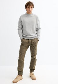 Suéter gris claro con un cuello alto, combinado con pantalones cargo oliva que cuentan con bolsillos laterales. Zapatillas beige completan el conjunto.