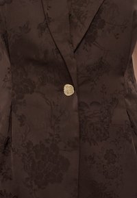 Σκούρο καφέ σακάκι με φλοράλ ζακάρ μοτίβο και ένα μοναδικό υφασμάτινο χρυσό κουμπί στο ύψος της μέσης.