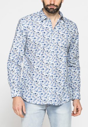 Uomo che indossa una camicia a maniche lunghe con bottoni, con motivo floreale blu, grigio e bianco e jeans azzurri.