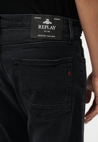 Κοντινή λήψη ενός ατόμου που φοράει μαύρο τζιν Replay, με εμφανή την πίσω τσέπη και την δερμάτινη ετικέτα με το λογότυπο πάνω από τη μέση.