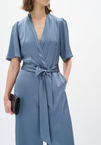 Hellblaues Wickelkleid mit kurzen Ärmeln, strukturiertem Stoff, V-Ausschnitt, passendem Gürtel, der an der Taille gebunden ist, und einer schwarzen Clutch in der Hand.