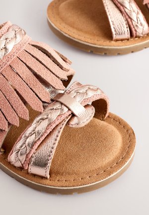 Sandales en daim rose avec des brides métalliques tressées et des détails à franges. Texture douce avec une semelle intérieure marron clair et une semelle extérieure en caoutchouc antidérapante.