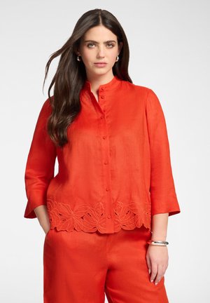 Pitkätukkainen nainen, joka käyttää kirkkaan oranssia nappilistallista paitapuseroa, jossa on kukkakuvioinen leikekoriste, ja vastaavia housuja, seisten käsi taskussa.