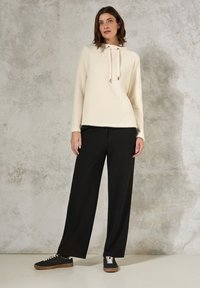 Pull crème texturé avec un col à cordon, associé à un pantalon noir ample et des baskets noires avec des lacets blancs et des semelles brunes.