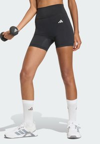 Pantaloncini sportivi neri con vita alta, caratterizzati da un design senza cuciture e un accento sottile. Indossati con calzini bianchi e scarpe sportive, mentre tiene un manubrio.