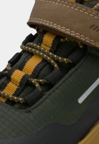 Gros plan sur une sneaker vert foncé avec une sangle marron, des lacets rayés jaunes et noirs, et un dessus en tissu texturé.