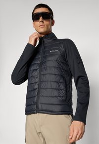 Man wearing black Columbia quilted jacket, beige pants, and large black sunglasses adjusting jacket collar against plain background.