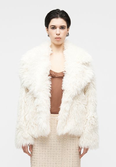Manteau en fausse fourrure blanche avec un col châle, doté d'une texture douce et volumineuse. Porté sur un haut en soie marron et une jupe beige ornée de perles.