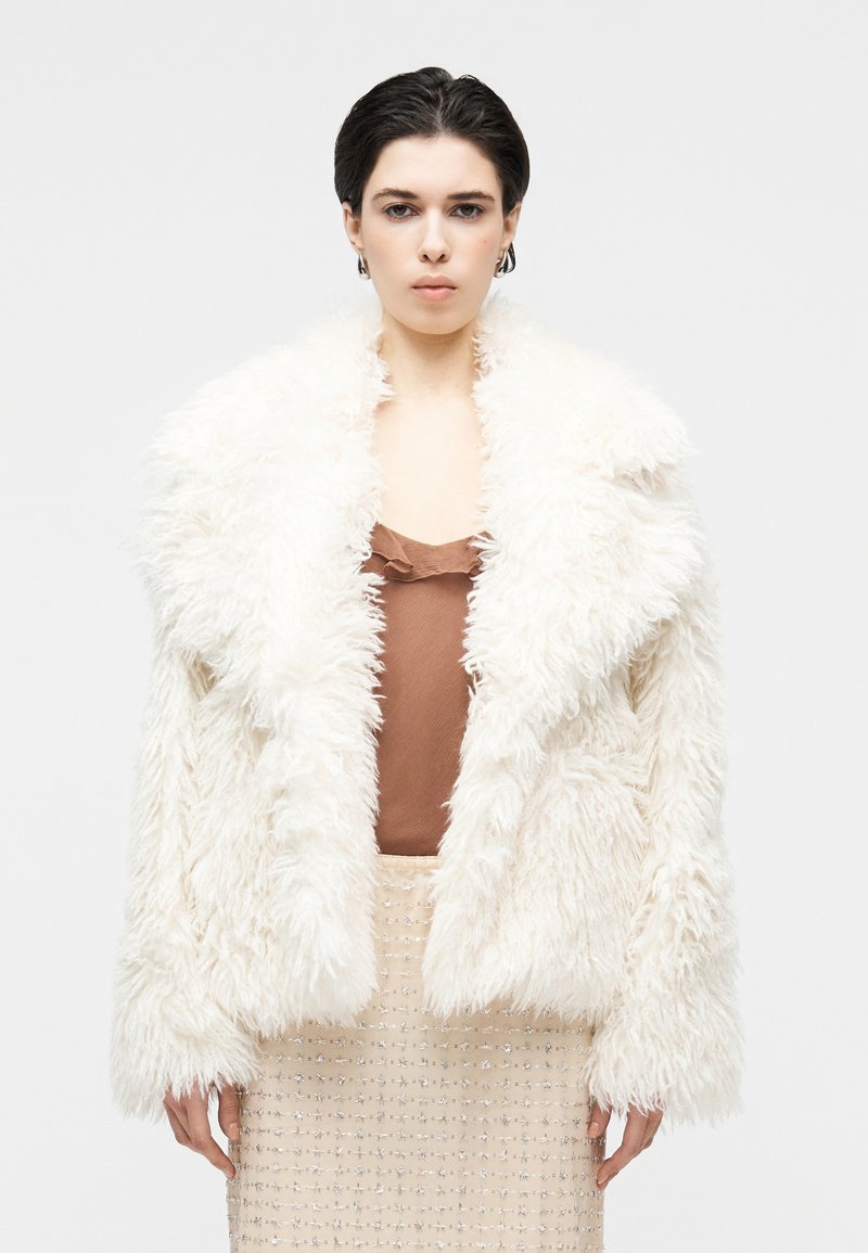 Bela umetna kožuh z ovratnikom šal, z razkošno teksturo. Nosi se čez rjavo svileno majico in bež krilo z biseri.