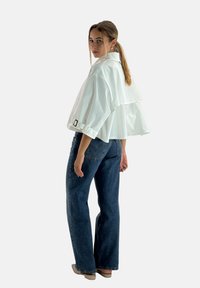 Hvid cropped jakke med lagdelt rygdesign, oprullede ærmer og sorte hardware-detaljer, der er parret med blå højtaljede jeans.