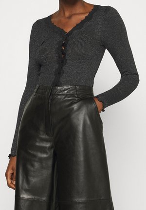 Frau trägt ein langärmliges, schwarzes, geripptes Oberteil mit Spitzenbesatz und Knöpfen, kombiniert mit schwarzen, hoch taillierten Leder-Hosen mit weitem Bein.