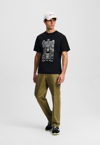 Juodas marškinėliai su baltu architektūriniu piešiniu ir tekstu, alyvinių spalvų kelnės ir šviesus raštuotas kepuraitė. Juodi sportbačiai su geltonais akcentais.