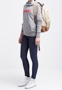 Felpa grigia con logo rosso "Bench.", tasca frontale, abbinata a jeans skinny scuri e sneakers alte bianche. Zaino a fantasia marrone e crema.