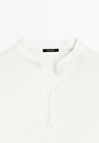 Hvid skjorte med krave med knaplukning og løkke. Glat stof med en subtil glans. Mærket lyder "Massimo Dutti, lavet i Marokko."