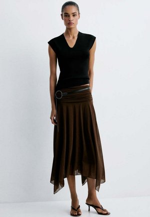 Femme portant un haut noir sans manches, une jupe asymétrique marron avec une ceinture noire et des sandales noires à talons, debout sur un fond blanc.