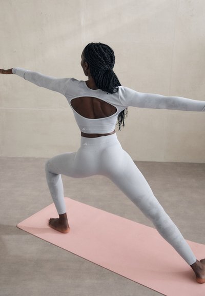 Top sportiv cu mânecă lungă de culoare gri deschis, cu un design cu spatele deschis, asortat cu colanți înalți pe talie, așezat pe o saltea de yoga roz.