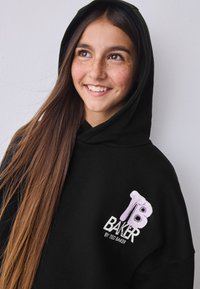 Felpa nera con cappuccio e un grande logo "TB" viola e bianco sul lato sinistro del petto. Tessuto liscio e design rilassato. Maniche lunghe e tasca a marsupio.