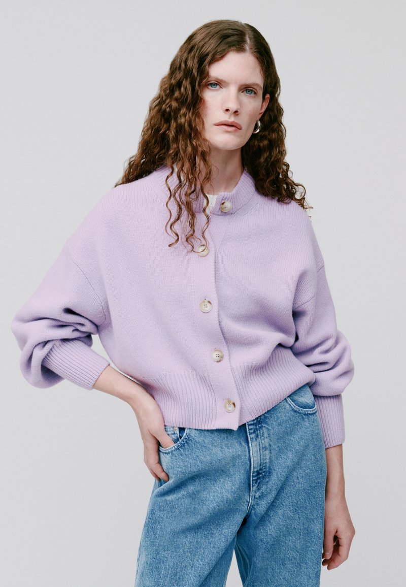 Lila gestrickte Strickjacke mit hohem Kragen und großen Knöpfen, mit lässigen Ärmeln und geripptem Saum. Kombiniert mit hellblauen Jeans.