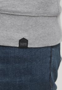 Grauer Hoodie mit geripptem Saum, versehen mit einem schwarzen sechseckigen Etikett, auf dem "CASUAL FRIDAY" steht. Über dunkelblauen Jeans getragen.
