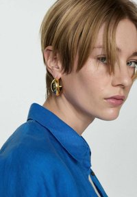 Orecchini a cerchio in oro con finitura lucida, che riflettono la luce. Il modello indossa una camicia blu con colletto, mettendo in evidenza i gioielli.