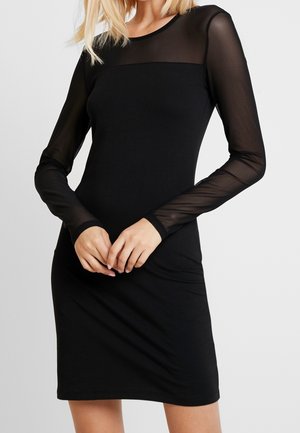 Mujer con un vestido negro ajustado con mangas largas transparentes y un panel superior transparente en el pecho sobre un fondo liso.