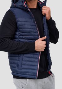 Smanicato imbottito blu navy con cappuccio, trama trapuntata, strisce di accento rosse e bianche all'interno del colletto, tasche laterali con zip e chiusura con cerniera.