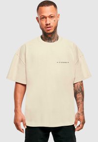 Bēša oversized t-kreklā ar īsām piedurknēm un apaļu kakla izgriezumu. Priekšpusē ir neliels pelēks logo. Audums izskatās mīksts.