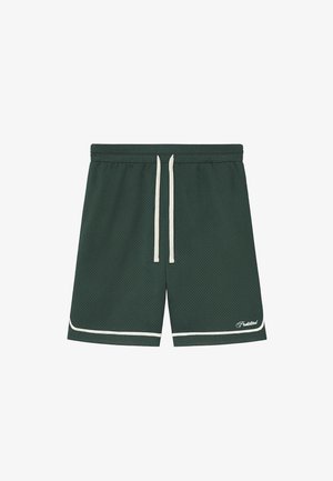 Męskie nogi ubrane w ciemnozielone sportowe szorty, białe skarpetki z napisem oraz brązowe mokasyny z zamszu na jasnoszarym tle.