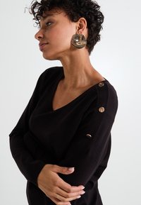 Suéter negro con cuello en V, adornado con botones metálicos en el hombro. La modelo lleva pendientes metálicos grandes y redondos. Textura de punto suave.