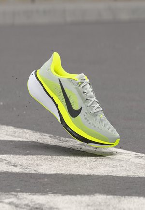 Sivá a neónovo žltá bežecká topánka s čiernym logom Nike, zachytená vo vzduchu nad spevneným povrchom s bielymi značeniami.