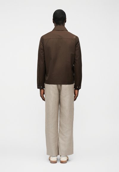 Άνδρας που στέκεται πλάτη, φοράει σκούρο καφέ μπουφάν, μπεζ παντελόνι με φαρδιά πόδια και λευκά αθλητικά παπούτσια σε απλό φόντο.