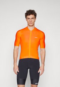 Krátky cyklistický dres z jasne oranžového materiálu, ktorý má predný zips a priliehavý strih. Spárované s čiernymi cyklistickými šortkami so pruhmi.