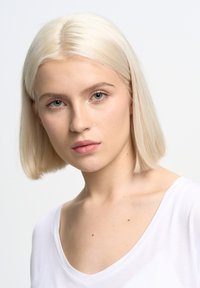 Blont hår i en rak bobfrisyr, ljus hy, klädd i en vit T-shirt med V-ringning. Klara blå ögon, minimal smink och ljusa fräknar synliga.