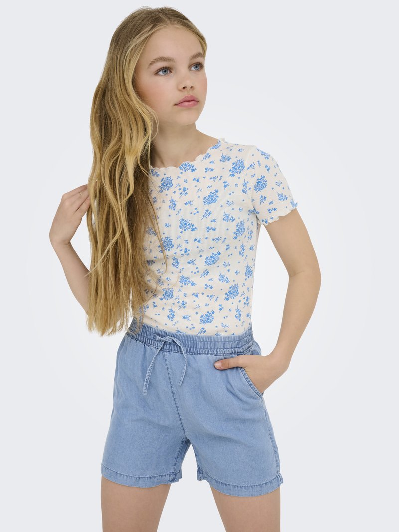Ragazza con lunghi capelli biondi che indossa una maglietta bianca a maniche corte con fiori blu e pantaloncini azzurri con coulisse, che guarda di lato.