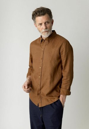 Middelbare man met grijze baard, gekleed in een bruin overhemd met knopen en een donkerblauwe broek, staand met één hand in de zak, kijkend naar opzij.