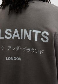 Mörkgrå sweatshirt med "ALLSAINTS" och "LONDON" i stora vita bokstäver, samt ytterligare text på japanska tecken. Slät bomullstextur.