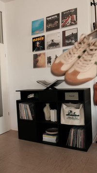 Białe i beżowe retro sneakersy wiszą nad czarną półką z winylowymi płytami, książkami, ozdobnym wazonem i torbą płócienną.