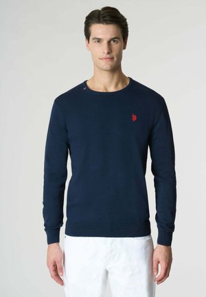 Uomo che indossa un maglione a maniche lunghe blu navy con un piccolo stemma rosso sul petto e pantaloni bianchi, in piedi contro uno sfondo chiaro e semplice.