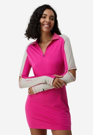 Donna sorridente in vestito rosa acceso con maniche lunghe beige con fori per i pollici e cerniera frontale, in piedi davanti a uno sfondo bianco semplice.