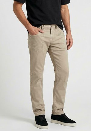 Man draagt beige rechte broek en zwarte sneakers, staat met één hand in de zak tegen een effen achtergrond.