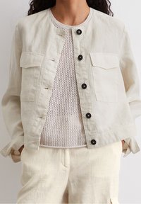 Lyse beige linnedjakke med rund halsudskæring, knaplukning fortil og to brystlommer; lagd over en tekstureret cremet striktrøje.