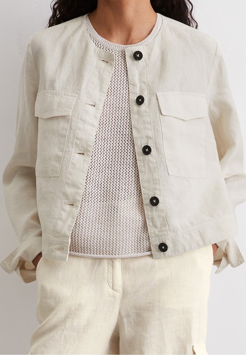 Lyse beige linnedjakke med rund halsudskæring, knaplukning fortil og to brystlommer; lagd over en tekstureret cremet striktrøje.