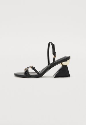Sandale noire à lanières avec une fine bride autour de la cheville, semelle plate et accent géométrique noir et or sur le talon. Présente des détails en métal.