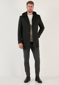 Abrigo negro texturizado con capucha, botones frontales, bolsillos laterales, combinado con jeans grises y botas de tobillo. Camisa interior blanca lisa visible.