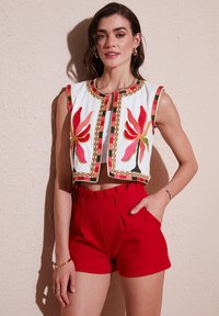 Gilet court brodé avec des motifs floraux rouges et jaunes, en tissu blanc, et bordures multicolores associé à un short rouge taille haute.