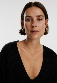 Zwarte gebreide trui met een V-hals ontwerp. Model draagt grote gouden en groene oorbellen. Glad huid met lichte sproeten.
