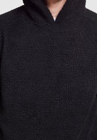 Felpa nera in pile con superficie texture, dotata di cappuccio e senza chiusure visibili. Il tessuto appare morbido e caldo.