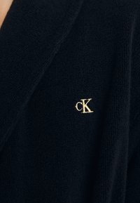 Jūras spēku krāsas auduma halāts ar mīkstu tekstūru, ar zelta izšuvumu "cK" logo. Vienkāršs dizains, bez rakstiem vai papildu akcentiem.