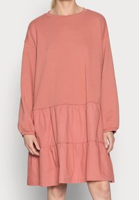 Vaaleanpunainen puuvillamekko, jossa on pitkät hihat, pyöreä pääntie ja kerrostettu röyhelöpohja. Pehmeä rakenne rentoa istuvuutta ja laskeutuvia hihoja.